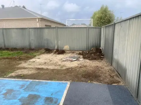 Bare soil and sand behind a grey metal fence, with a blue-painted concrete patch and dark paving at the bottom