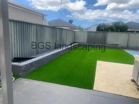 Artificial grass edged by a low dark block wall along a grey metal fence, with dark paving and a pale concrete patch in the foreground