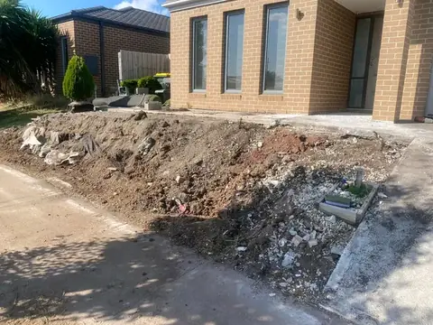 Cleared ground with soil and rubble beside a concrete driveway, in front of a tan brick house