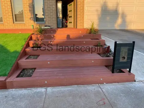 Close-up of red-brown deck steps with a wood-grain texture, planter boxes and artificial grass at the left
