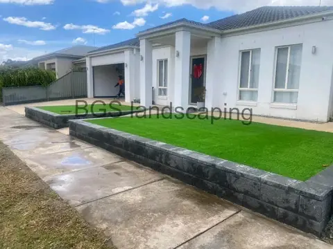 Long raised dark grey block bed with artificial grass along a concrete path, in front of a white house