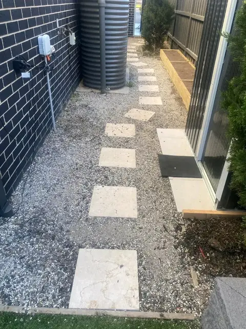 Gravel passage with large pale pavers laid as stepping stones beside a dark brick wall and a tall corrugated tank