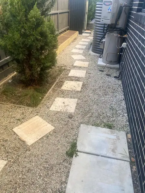 Gravel passage with pale pavers beside a timber fence, a conifer and air-conditioning units