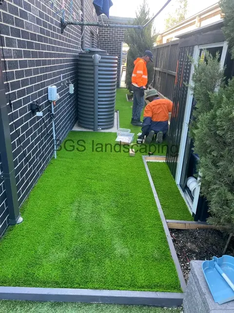 Artificial grass along a dark brick wall and a tall corrugated tank, with two workers in orange hi-vis