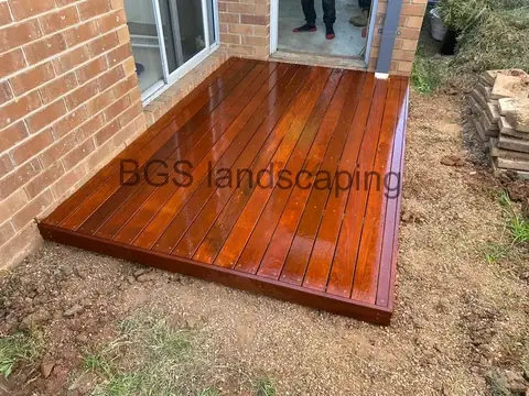 Glossy reddish-brown timber deck laid on bare ground against a brick wall, with stacked timber at the right